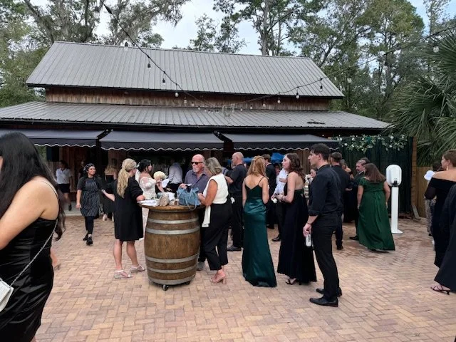 Group of people gathering outdoors in formal attire, standing around a large barrel table on a brick patio in front of a rustic building with string lights overhead and surrounded by trees.