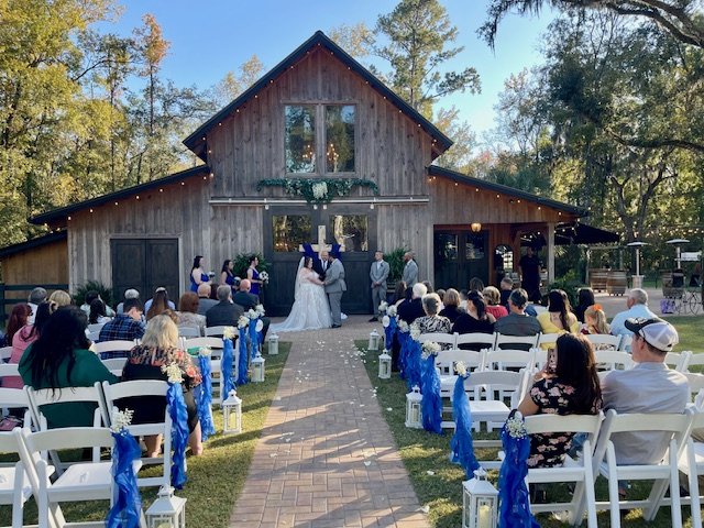 Outdoor wedding ceremony with a wooden barn, bride and groom, officiant, guests seated in white chairs with blue decorations, trees in the background.