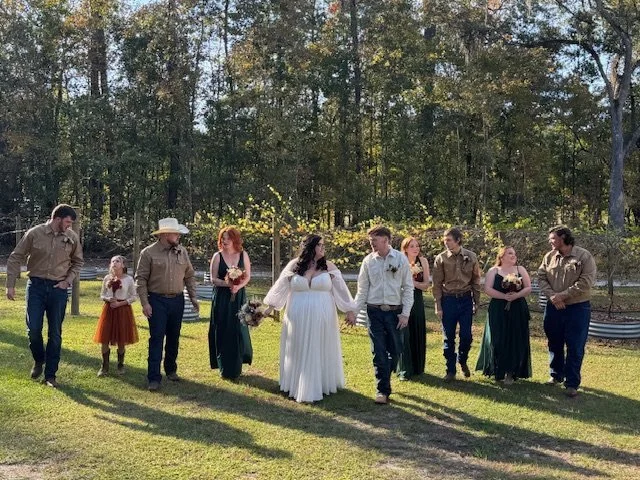 A wedding party walking outdoors on grass, with trees in the background. The bride is in a white gown holding a bouquet, flanked by bridesmaids in dark dresses and groomsmen in western-style shirts and jeans. A young girl in a red dress is on the lef