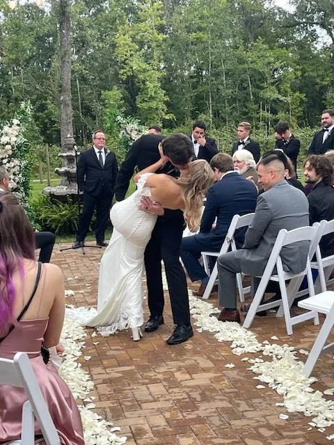 A wedding ceremony outdoors with a bride and groom sharing a kiss, surrounded by seated guests and friends, amidst a wooded setting decorated with flower petals.