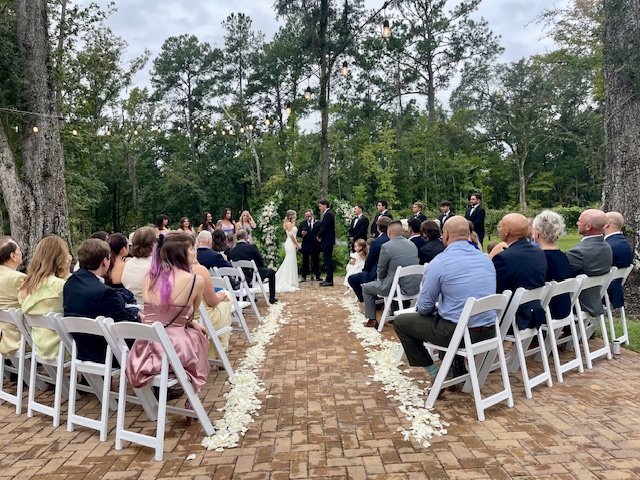 Outdoor wedding ceremony with guests seated on white chairs facing the couple, who are standing at the end of a brick path decorated with white flower petals, surrounded by trees and string lights hanging overhead.