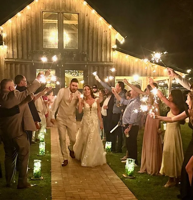 A bride and groom walking down an outdoor aisle at night, surrounded by friends and family holding sparklers in celebration.