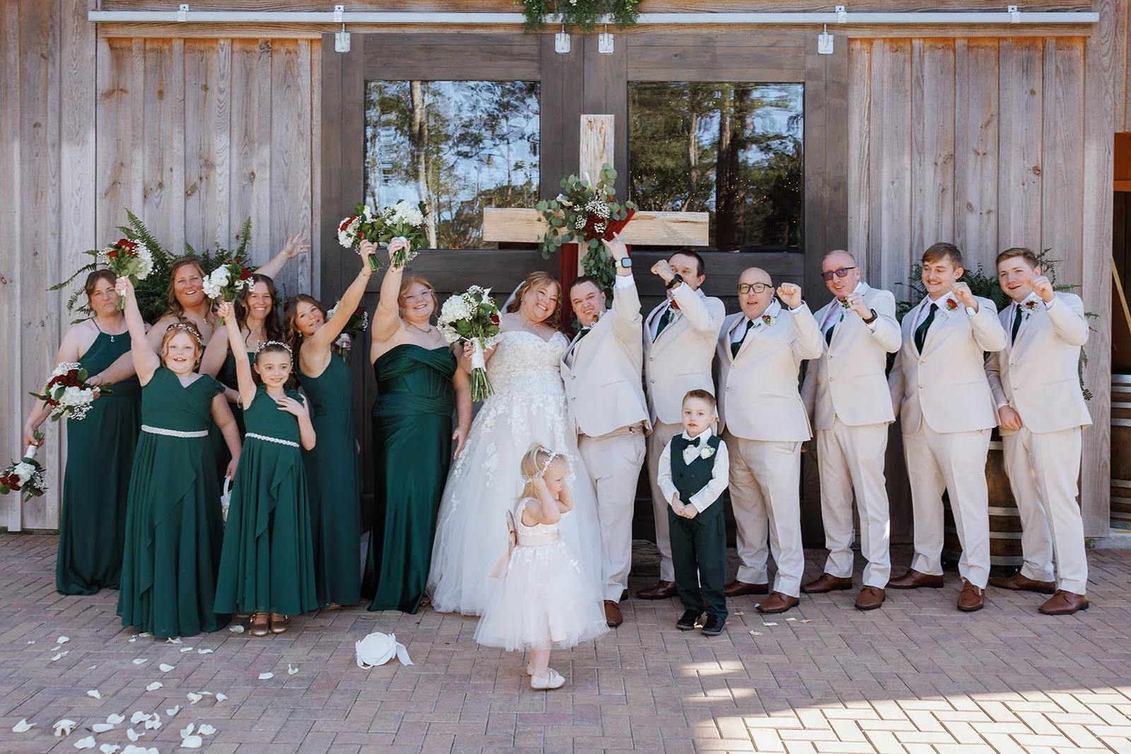 Group of people celebrating wedding in front of a building with a wooden exterior, including bride and groom in the center, bridesmaids in green dresses, groomsmen in white suits, children in formal attire, with flowers and confetti on the ground.