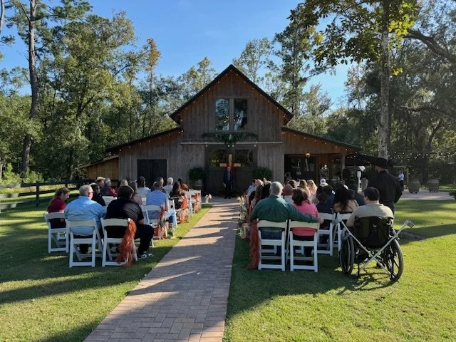 Outdoor wedding ceremony with guests seated on white chairs on either side of a brick pathway leading to a wooden chapel surrounded by trees on a sunny day.
