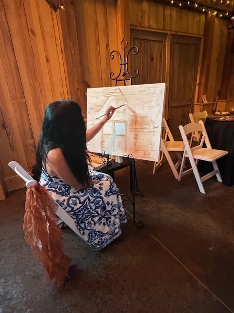 A woman with long dark hair painting on a canvas set up on an easel in a wooden room with string lights, sitting on a white folding chair.