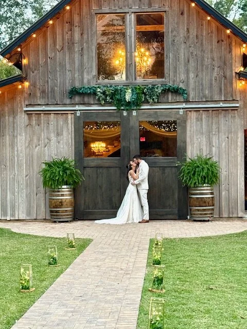A wedding couple sharing an intimate moment in front of a rustic wooden barn decorated with greenery and warm string lights, surrounded by potted plants and candle lanterns along a stone pathway.