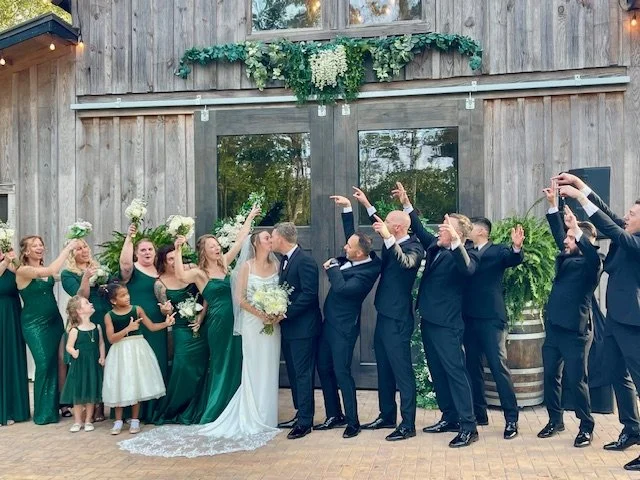 Bridal party and wedding guests standing outside a rustic barn, celebrating the wedding ceremony. The bride and groom are kissing, surrounded by bridesmaids in green dresses holding flowers, and groomsmen in suits, with some guests raising hands in c