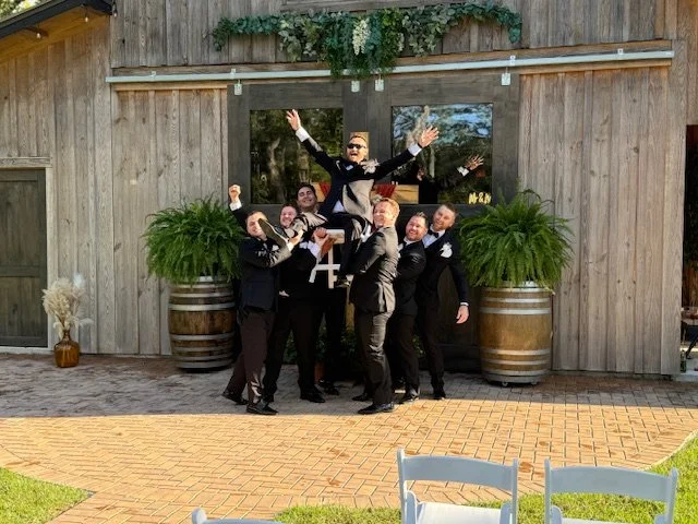 A group of men in black tuxedos lifting a man in a tuxedo in the air in front of a rustic wooden building with large potted plants on either side, outdoor setting.