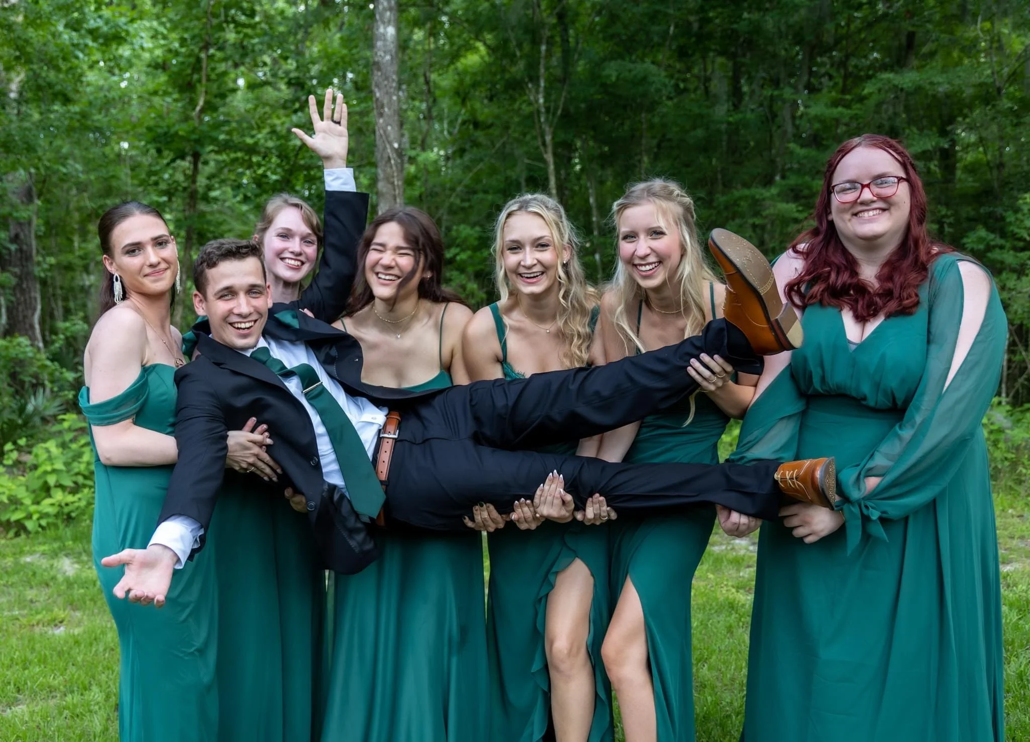 Group of seven young adults dressed in green dresses and a black suit with a green tie, standing outside in a green wooded area, smiling and celebrating together.