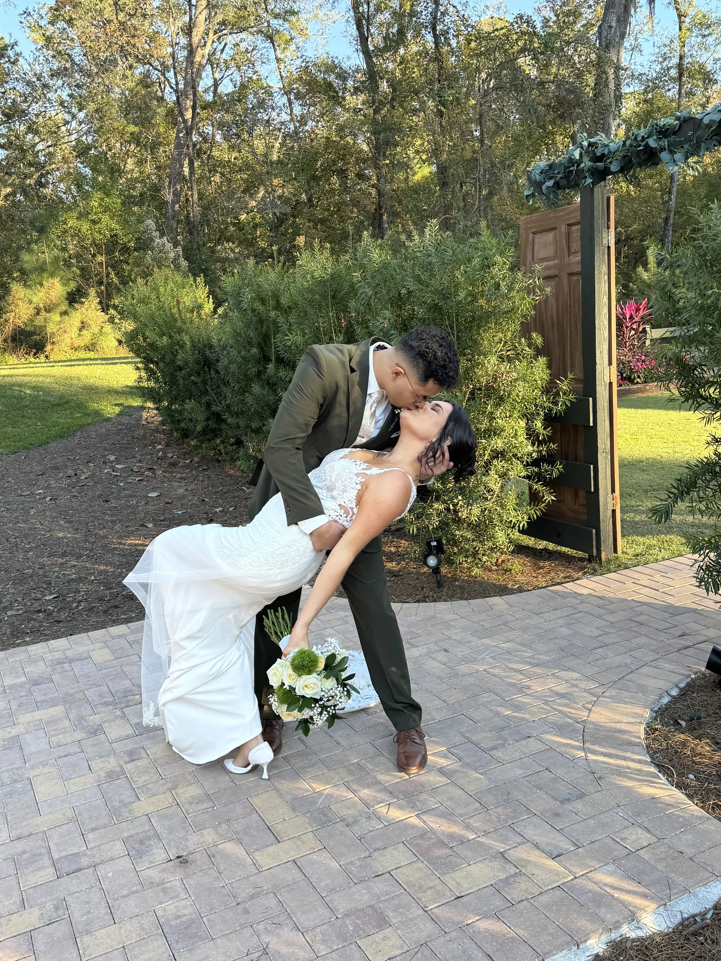 A newlywed couple is kissing outdoors, with the groom dipping the bride and holding her waist. The bride is holding a bouquet of white flowers, and they are standing on a paved pathway near a garden with green bushes and trees, under a clear sky.