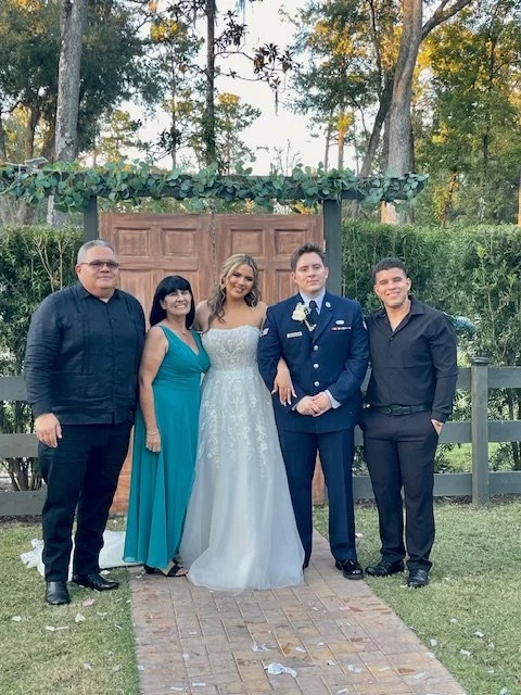 Group of five people standing outdoors in front of a wooden entrance with greenery. One woman in a white wedding gown and a man in a U.S. Air Force uniform are in the center, with a man and a woman on each side.