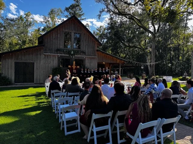 Outdoor wedding ceremony with guests seated on white chairs, a wooden barn in the background, and string lights hanging overhead during daytime.