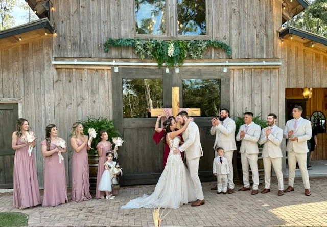 Bride and groom kissing in front of a rustic wooden barn during their wedding ceremony, with bridesmaids and groomsmen clapping and celebrating.
