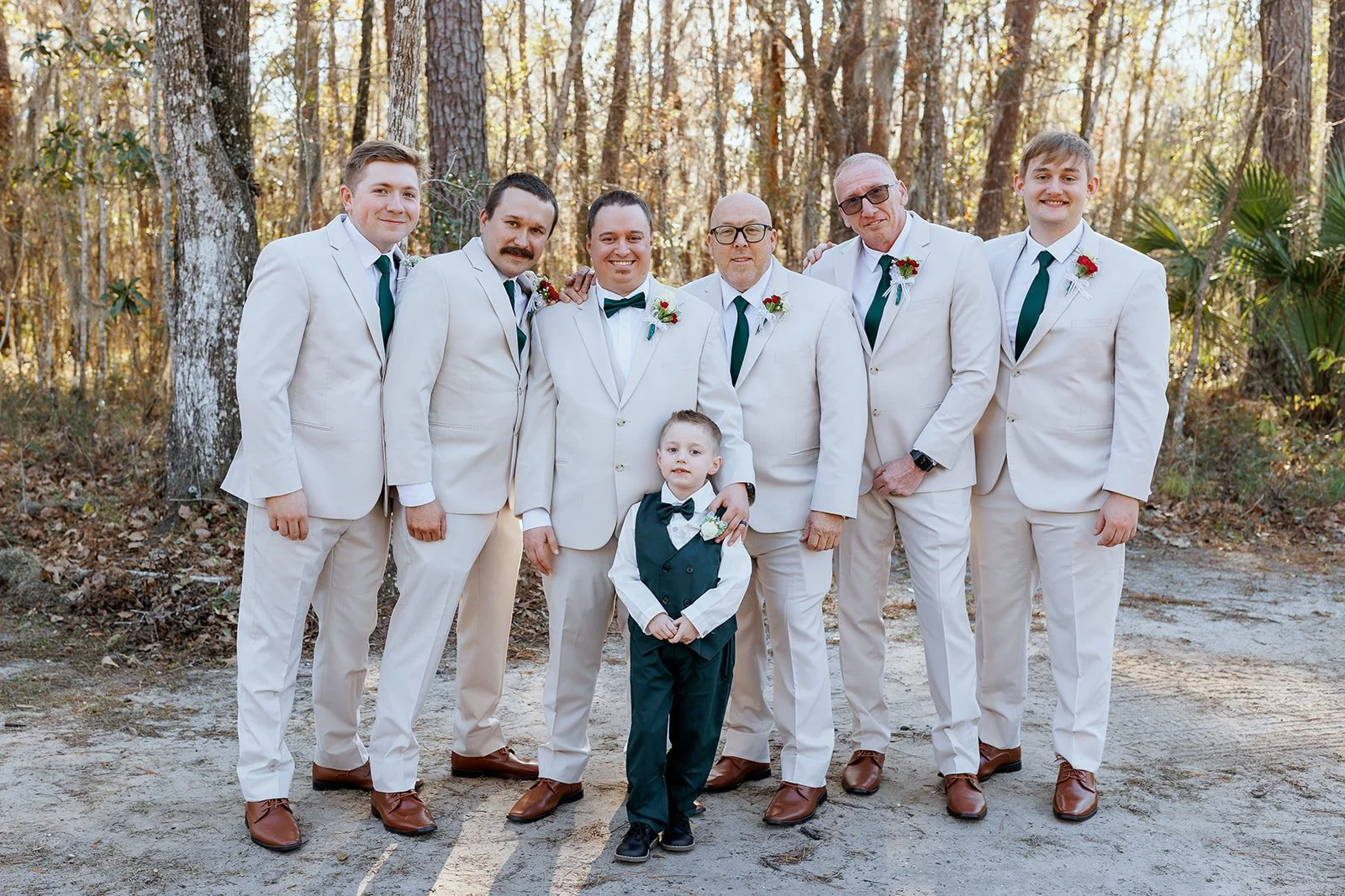 Group of men and a young boy dressed in white suits with green ties, standing outdoors in a wooded area during daytime.