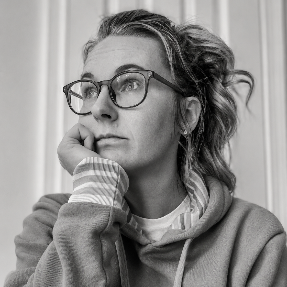 A black and white photo of a woman with glasses, resting her chin on her hand, looking thoughtful. She has wavy hair and is wearing a hoodie.
