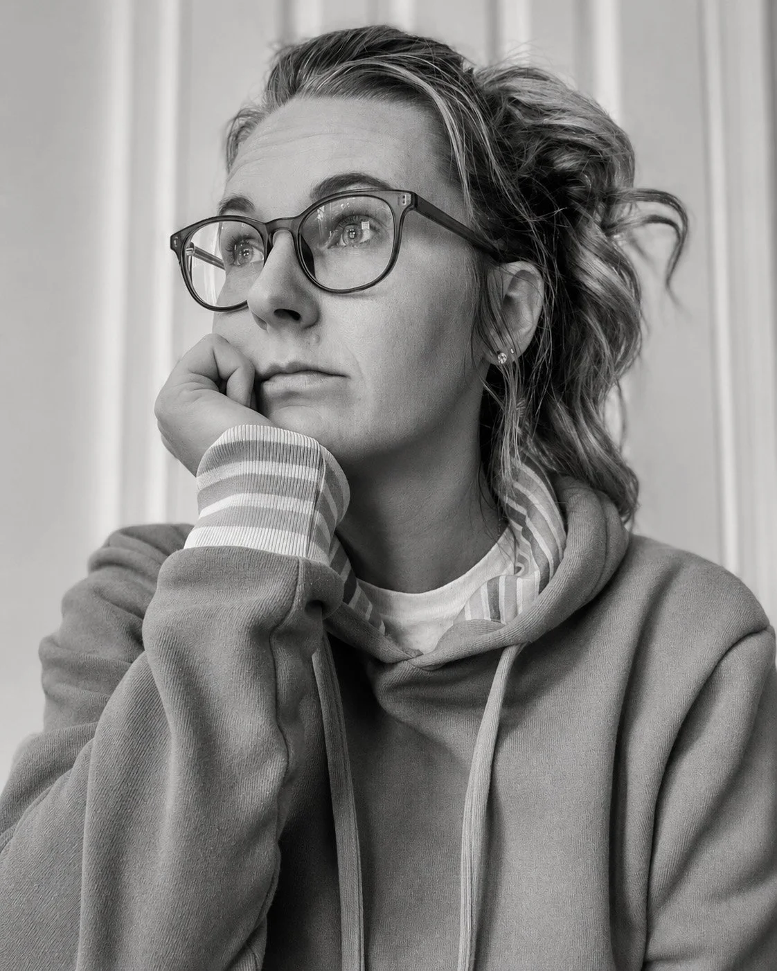A woman with wavy hair, glasses, and earrings, wearing a hoodie, resting her chin on her hand, looking thoughtfully to the side in a black and white photo.