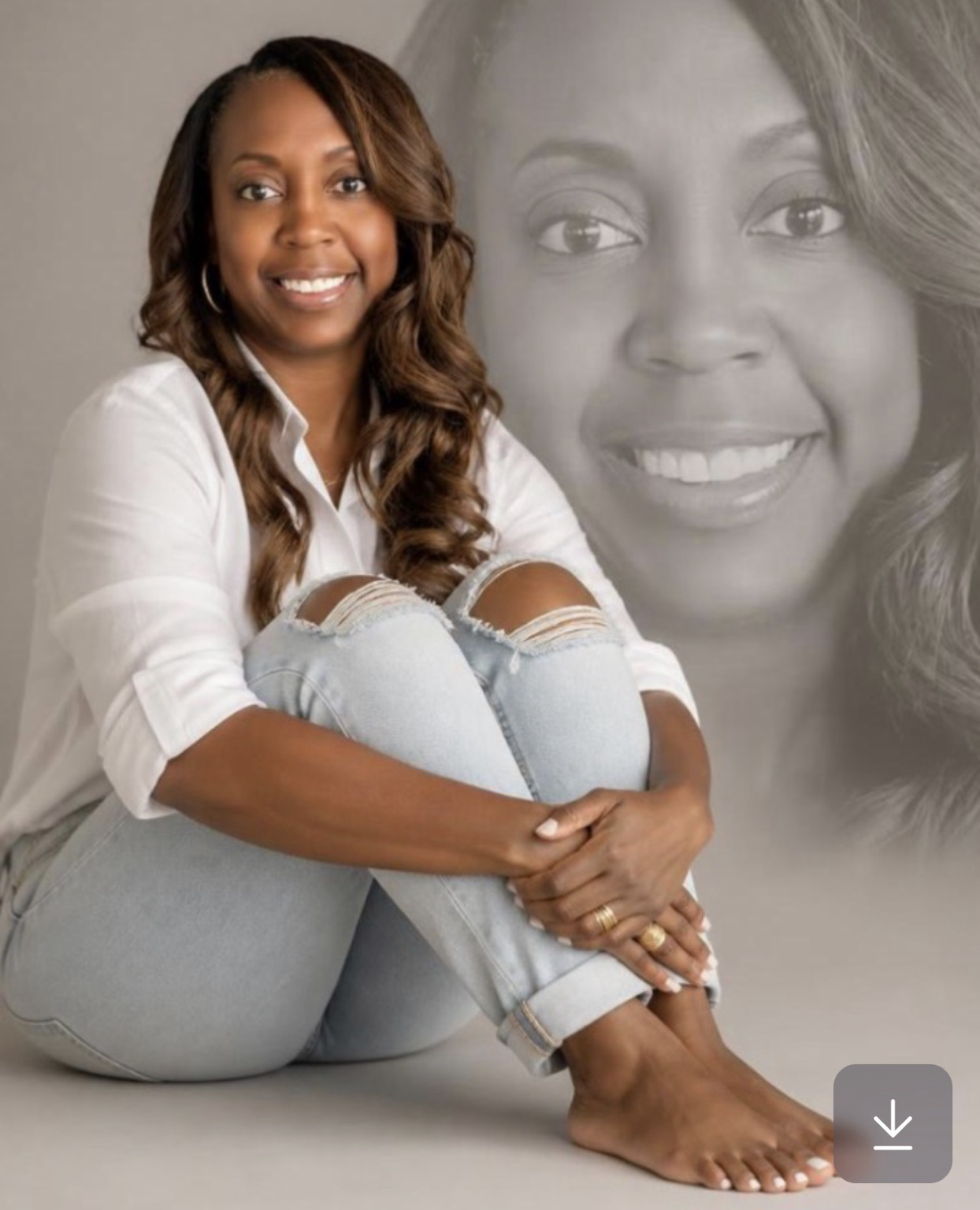 A smiling woman with long wavy brown hair, wearing a white shirt and ripped jeans, sitting cross-legged with her arms around her legs, in front of a large grayscale portrait of herself.