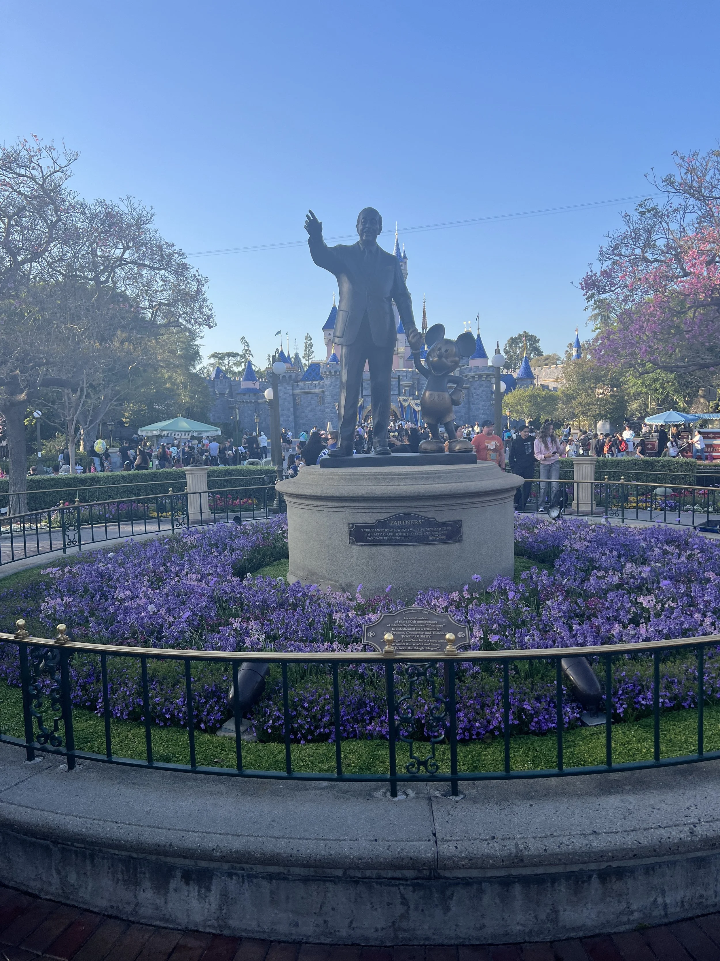 Statue of Walt Disney with Mickey Mouse in front of a castle at Disneyland, surrounded by purple flowers and a crowd of visitors.