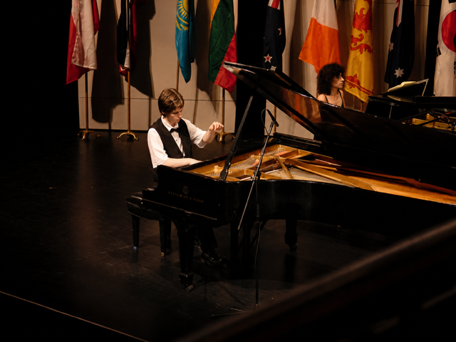 A young boy playing a grand piano on stage during a performance, with flags from various countries hanging in the background.