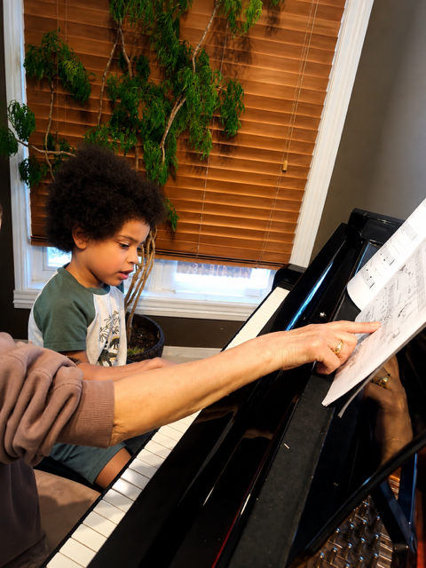A young boy and an adult practicing piano together, with the adult pointing at sheet music on the piano.