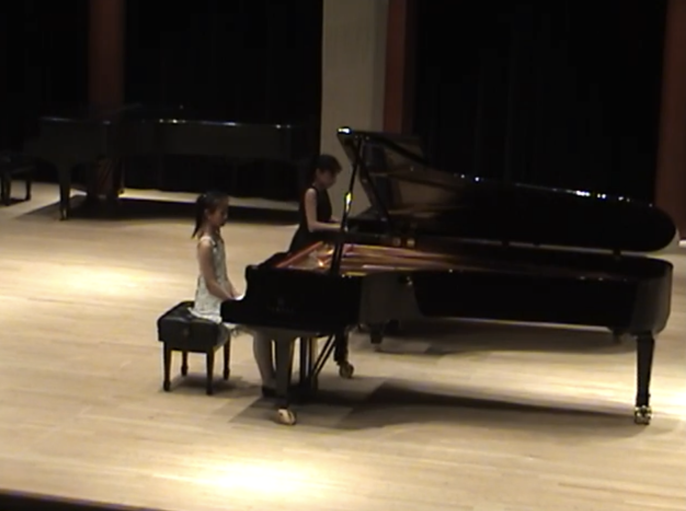 Two young girls practicing piano on a grand piano in a concert hall.