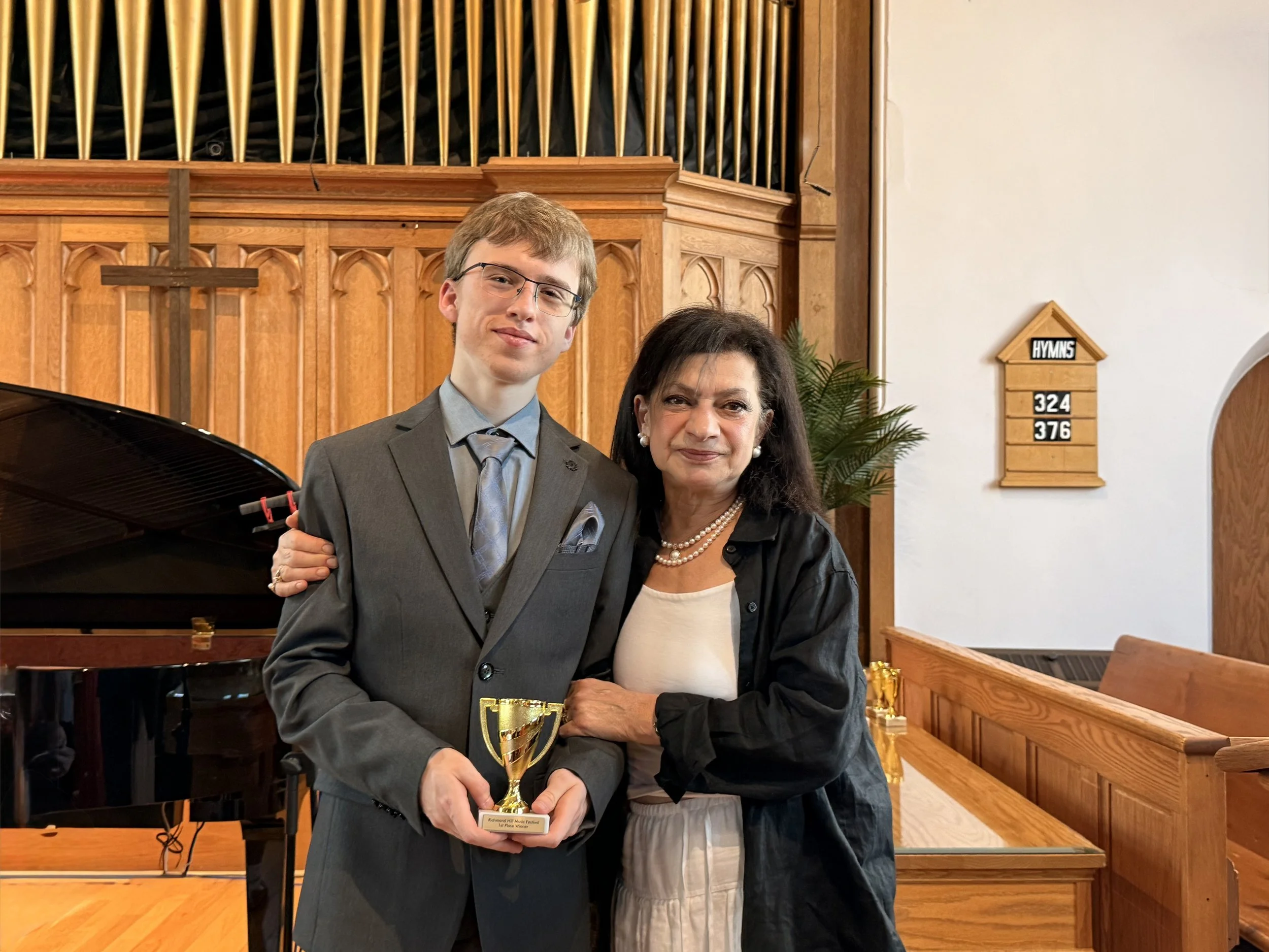 A young man in a gray suit and glasses and a woman in a black jacket and pearl necklace standing inside a church, holding a trophy. Behind them, a wooden cross and hymnal board are visible, with a grand piano partially seen in the foreground.