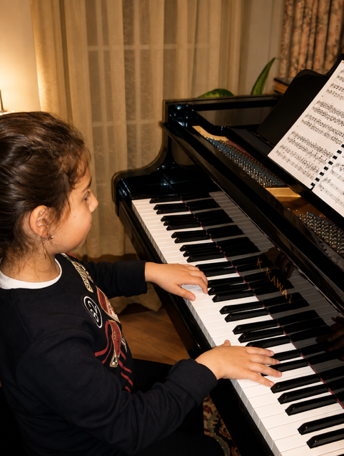 A young girl playing a black grand piano in a warmly lit room, with sheet music on the music stand.