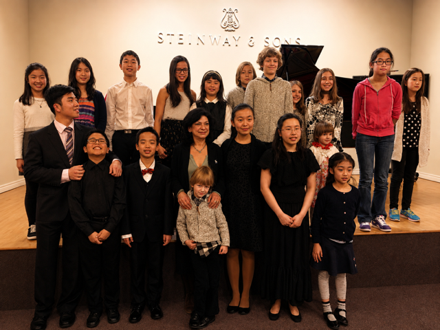 Group of children and an adult woman posing on a stage in a music studio, with a Steinway & Sons piano behind them.