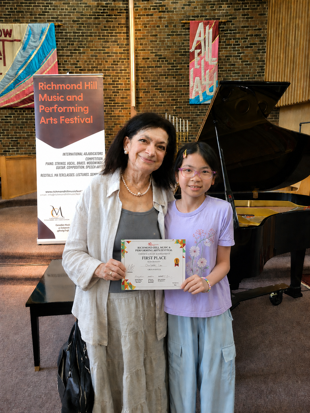 A woman and a girl with glasses standing together on stage, holding a first-place award certificate at the Richmond Hill Music and Performing Arts Festival, with a grand piano and festival banners in the background.