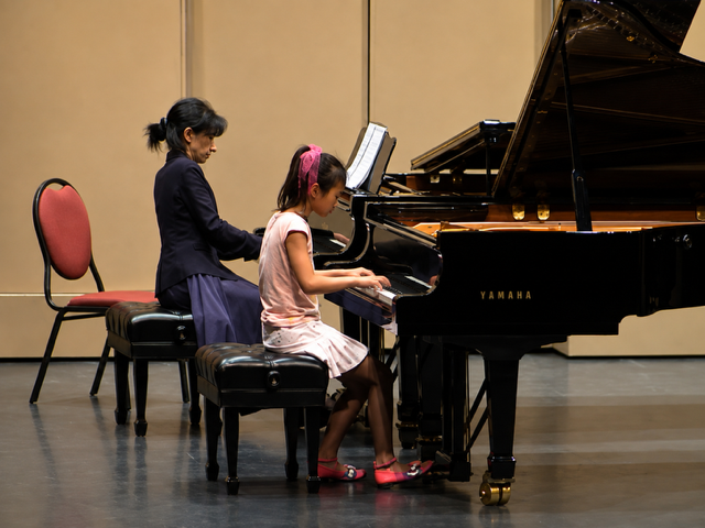 A woman and a young girl playing a Yamaha grand piano on a stage in a music rehearsal or recital setting.