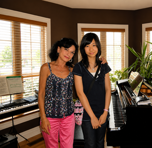 Two women, one older and one younger, standing together in a music room with a piano and a keyboard, both smiling and posing for the photo.