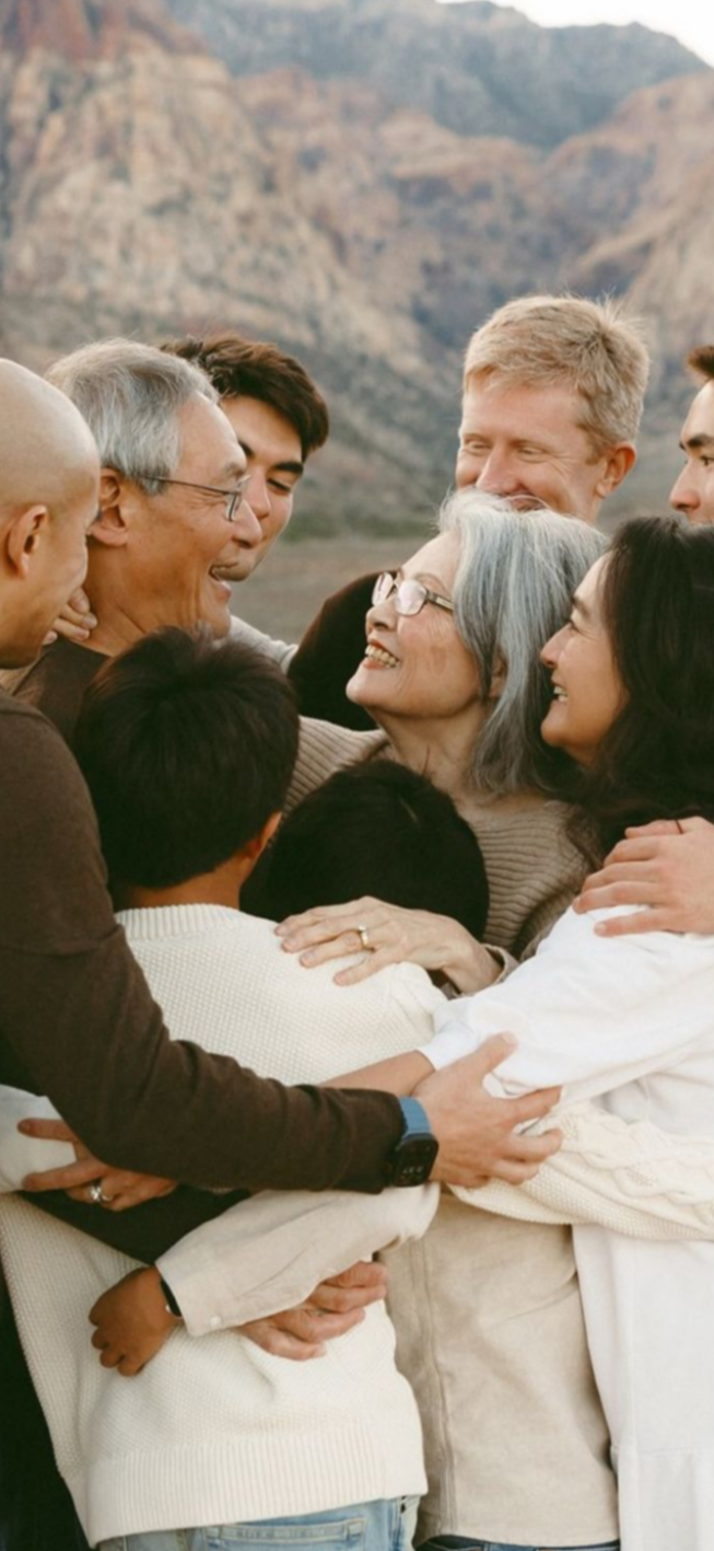 A diverse group of people, including young children, adults, and elderly, hugging and smiling together outdoors with mountains in the background.