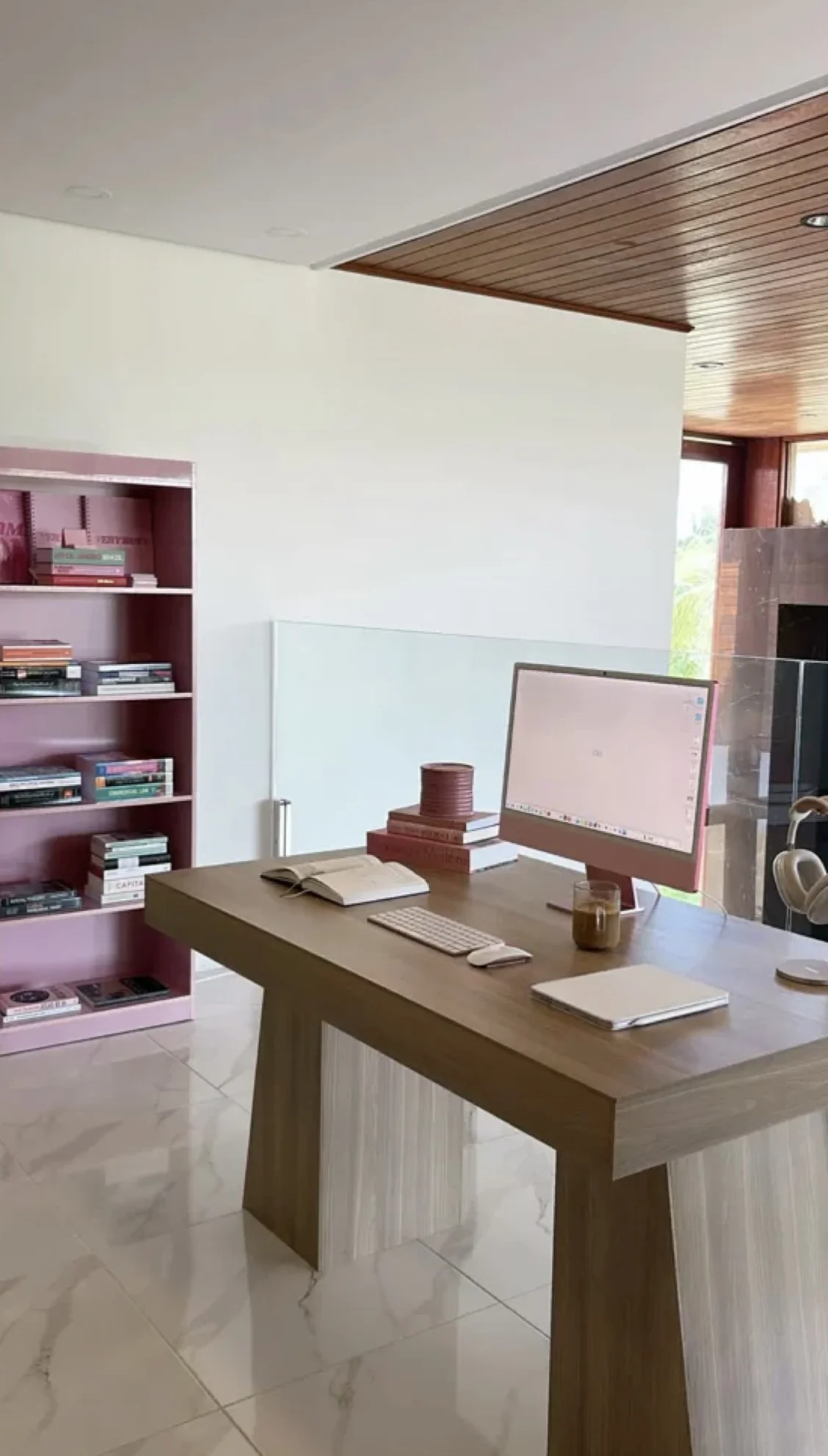 A modern home office with a wooden desk, pink bookshelf filled with books, a computer monitor, open notebook, mouse, keyboard, and a cup of coffee. There is a ceiling with wooden paneling and large windows in the background.