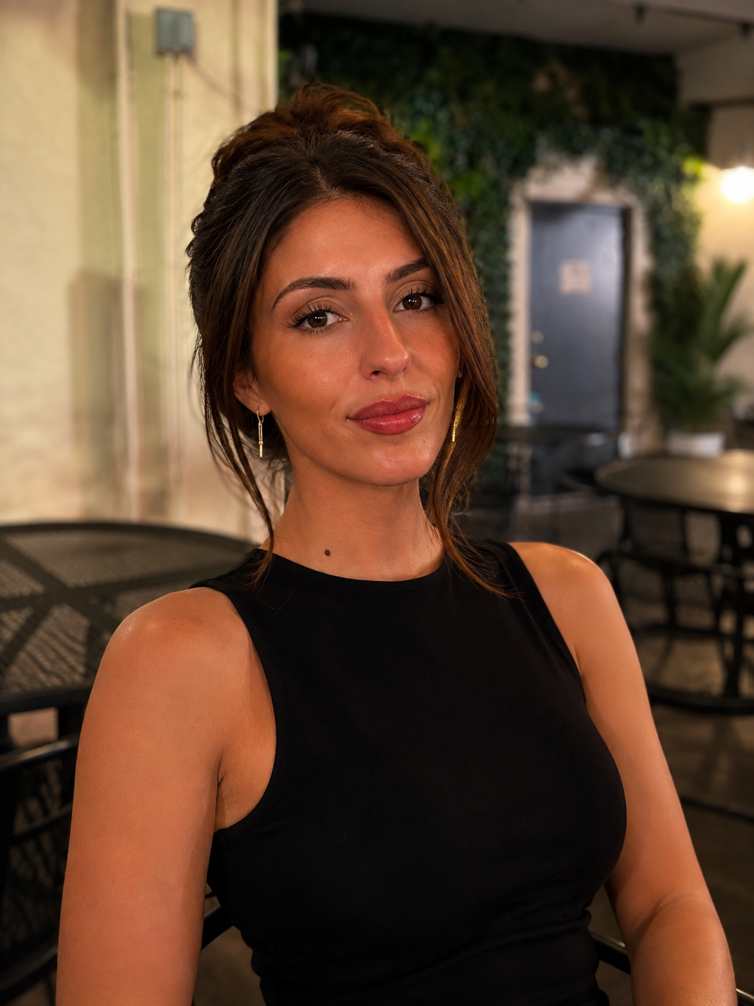 A woman with shoulder-length brown hair and earrings, wearing a black sleeveless top, sitting at an outdoor cafe with tables and chairs, green plants, and a doorway in the background.