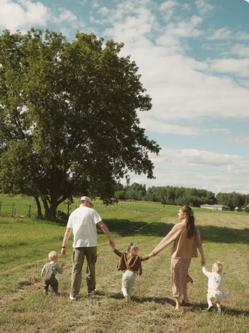 A family walks hand-in-hand on a grassy field near a large tree under a partly cloudy sky. Two adults and three young children are holding hands and walking together.
