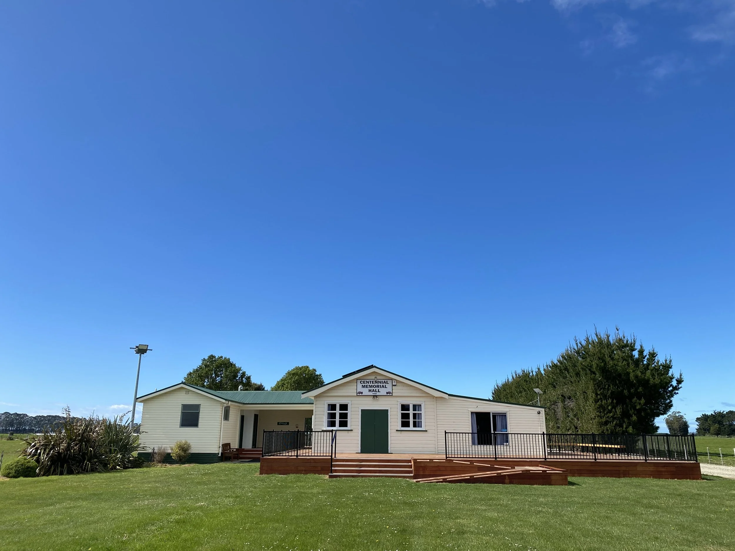 A small white building labeled 'Centennial Memorial Hall' with a green door and windows, surrounded by a grassy lawn, bushes, trees, and a clear blue sky.