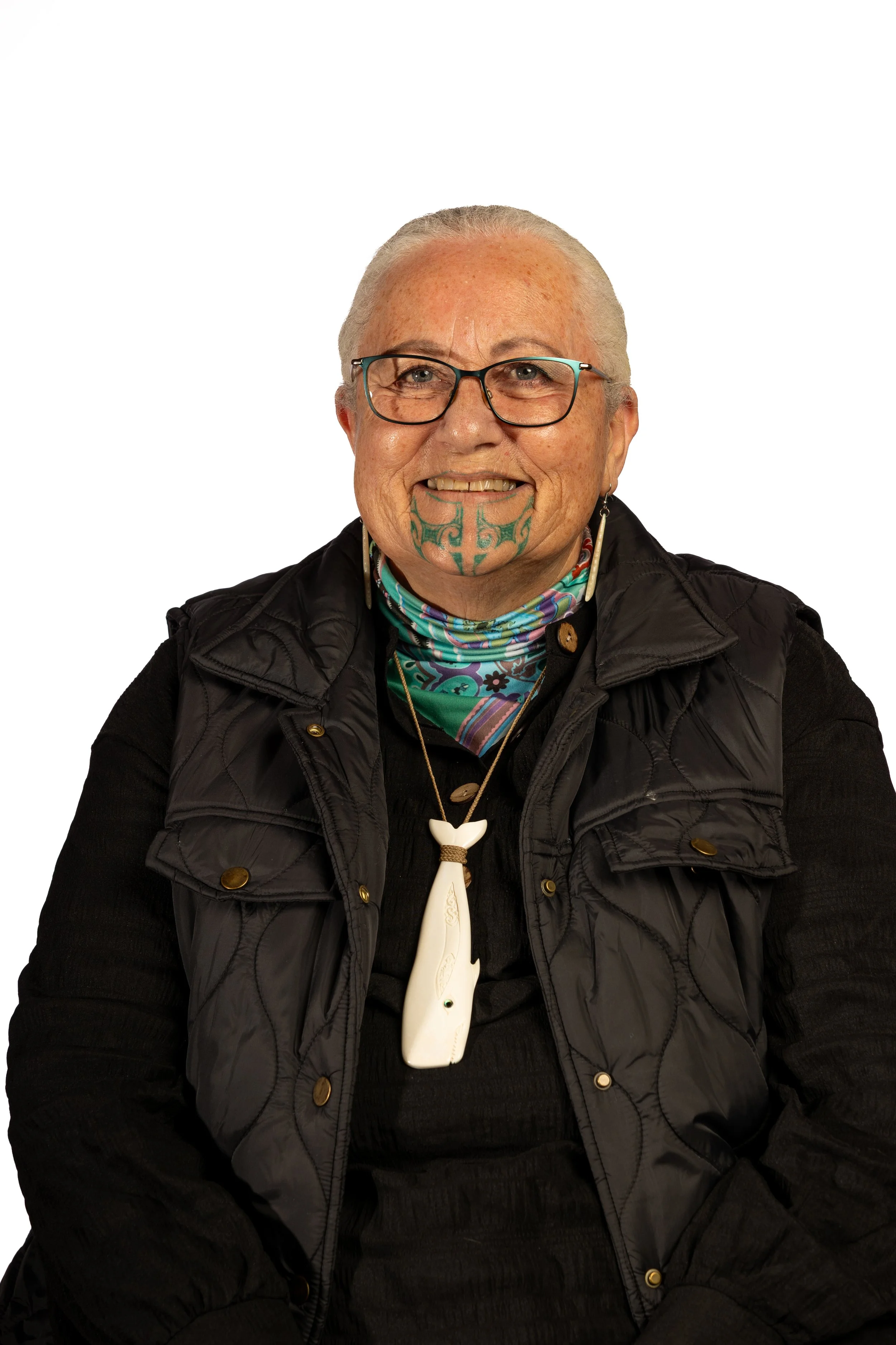 An elderly woman with glasses and a large Maori chin tattoo smiling, wearing a black vest, colorful scarf, and a whale-shaped necklace.
