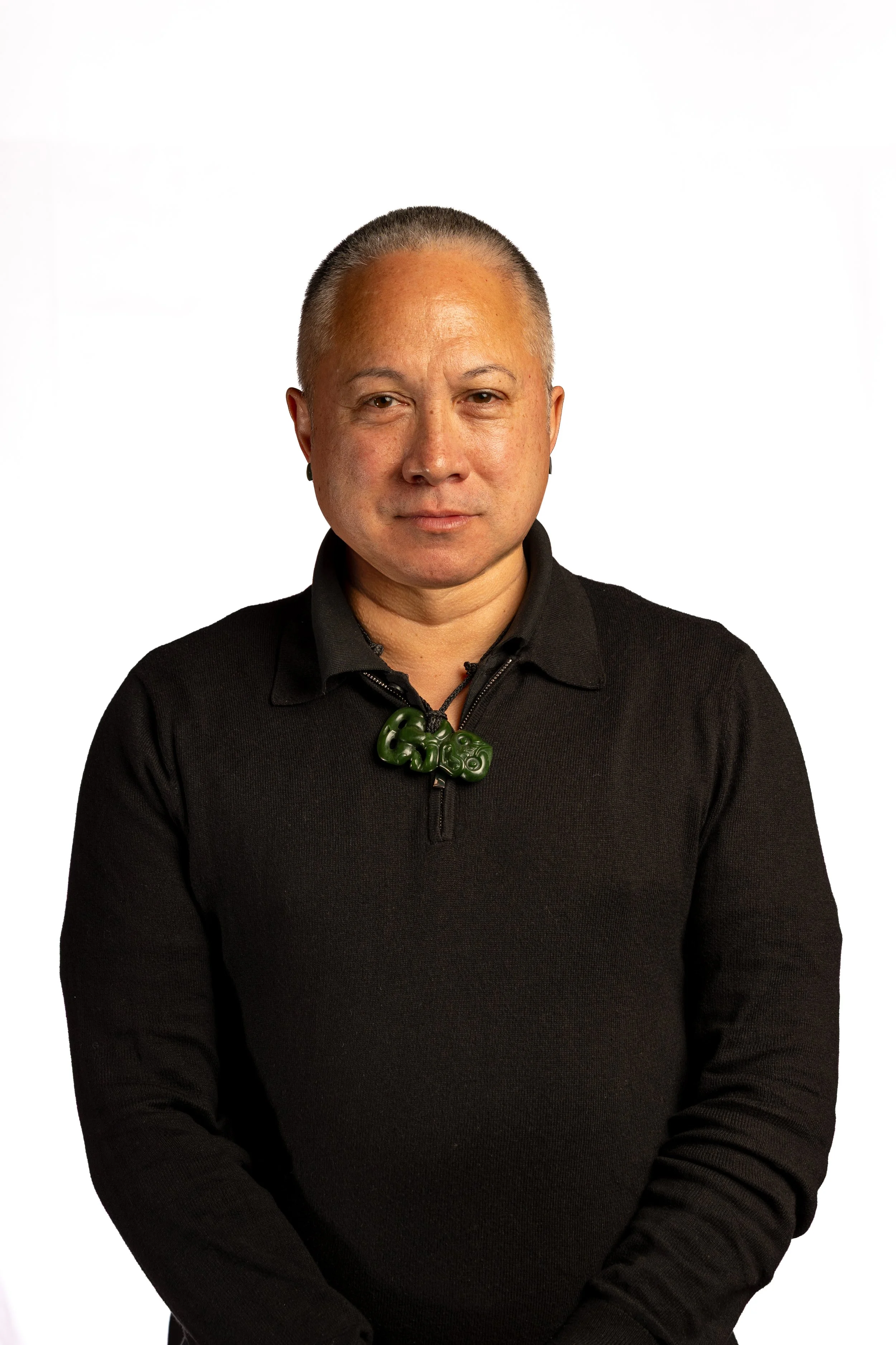 Portrait of a man with short gray hair, wearing a black zip-up sweater with a green jade necklace, standing against a plain white background.