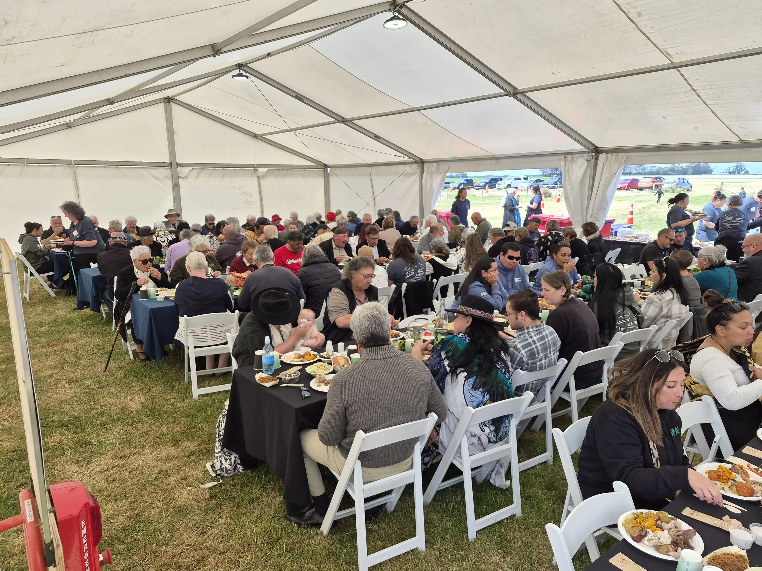 A large outdoor event tent filled with people sitting at tables, eating food, and socializing. Outside the tent, cars are parked on the grass.