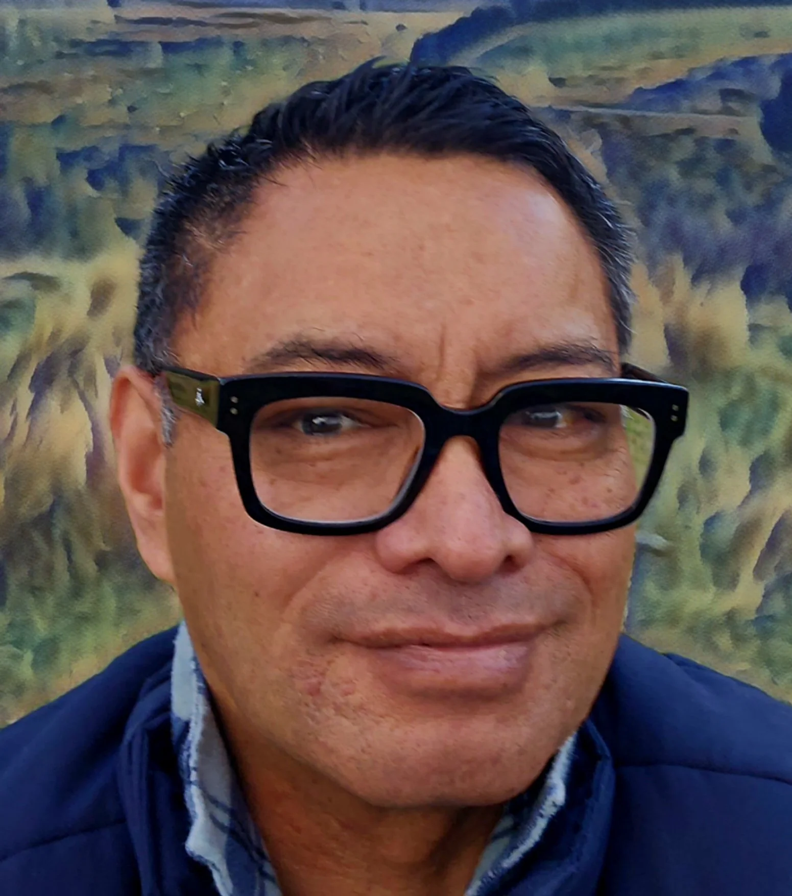 Close-up of a man with black hair, wearing black glasses, and a blue jacket, standing in front of a landscape with hills and trees.