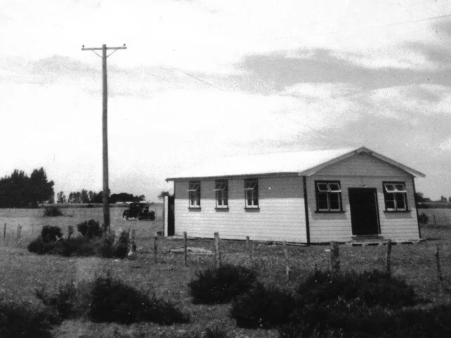 Black and white photo of a house with a gable roof, multiple windows, and a door, situated in a rural landscape with tall grass and sparse bushes. A utility pole is in the foreground, and a car is seen in the background.