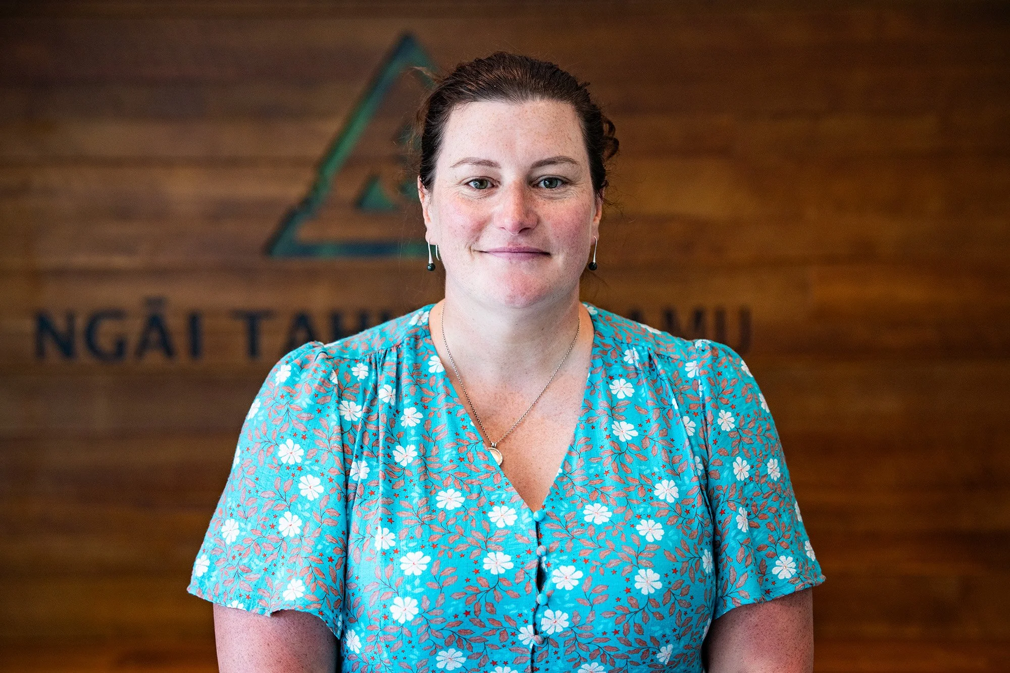 A woman with dark hair tied back, wearing a blue floral dress, standing in front of a wooden wall with the text 'Ngai Tahu' partially visible behind her.