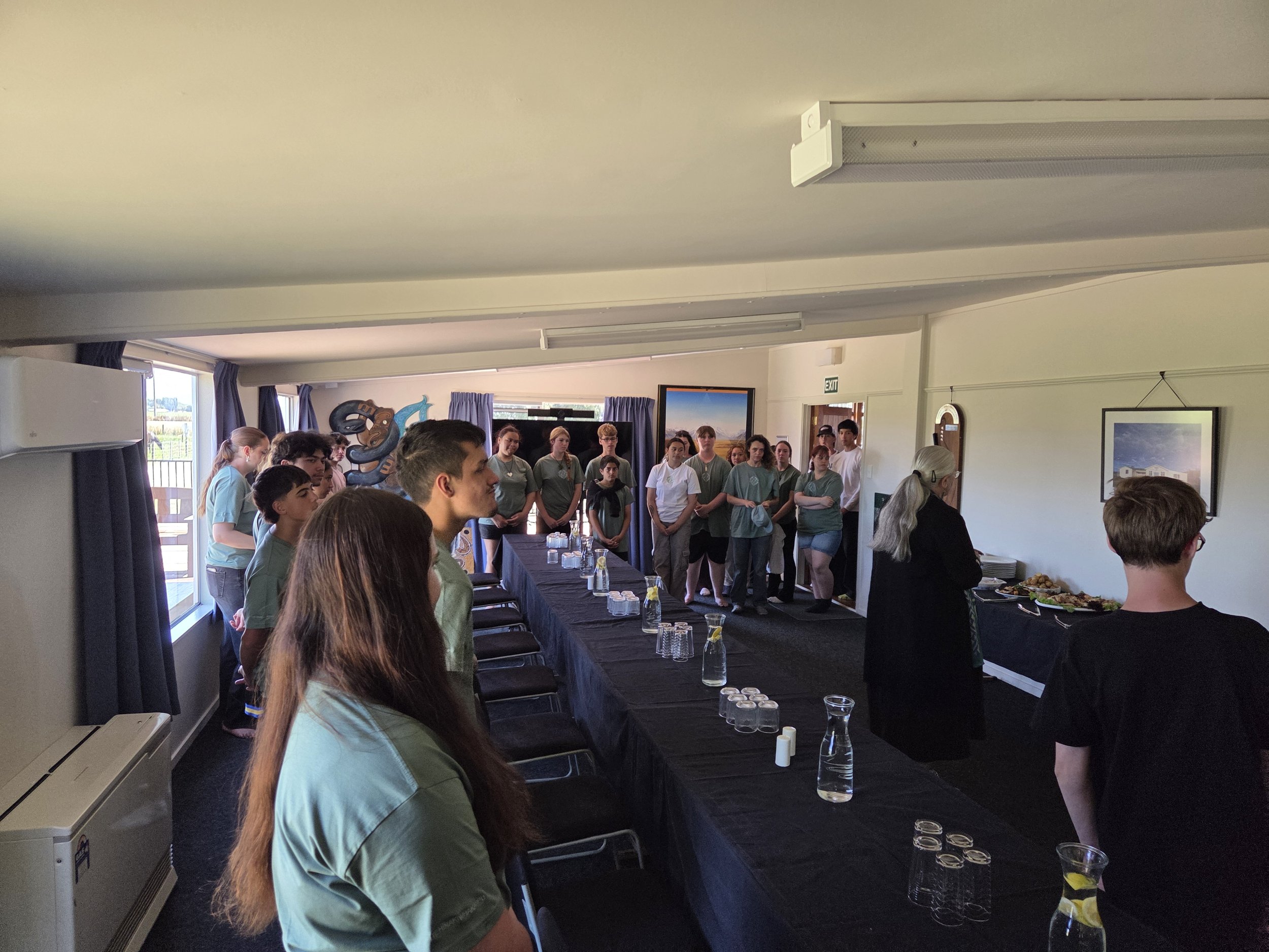 People standing in a room with a table set with water bottles and glasses, with some food towards the back of the room, during a gathering or meeting.