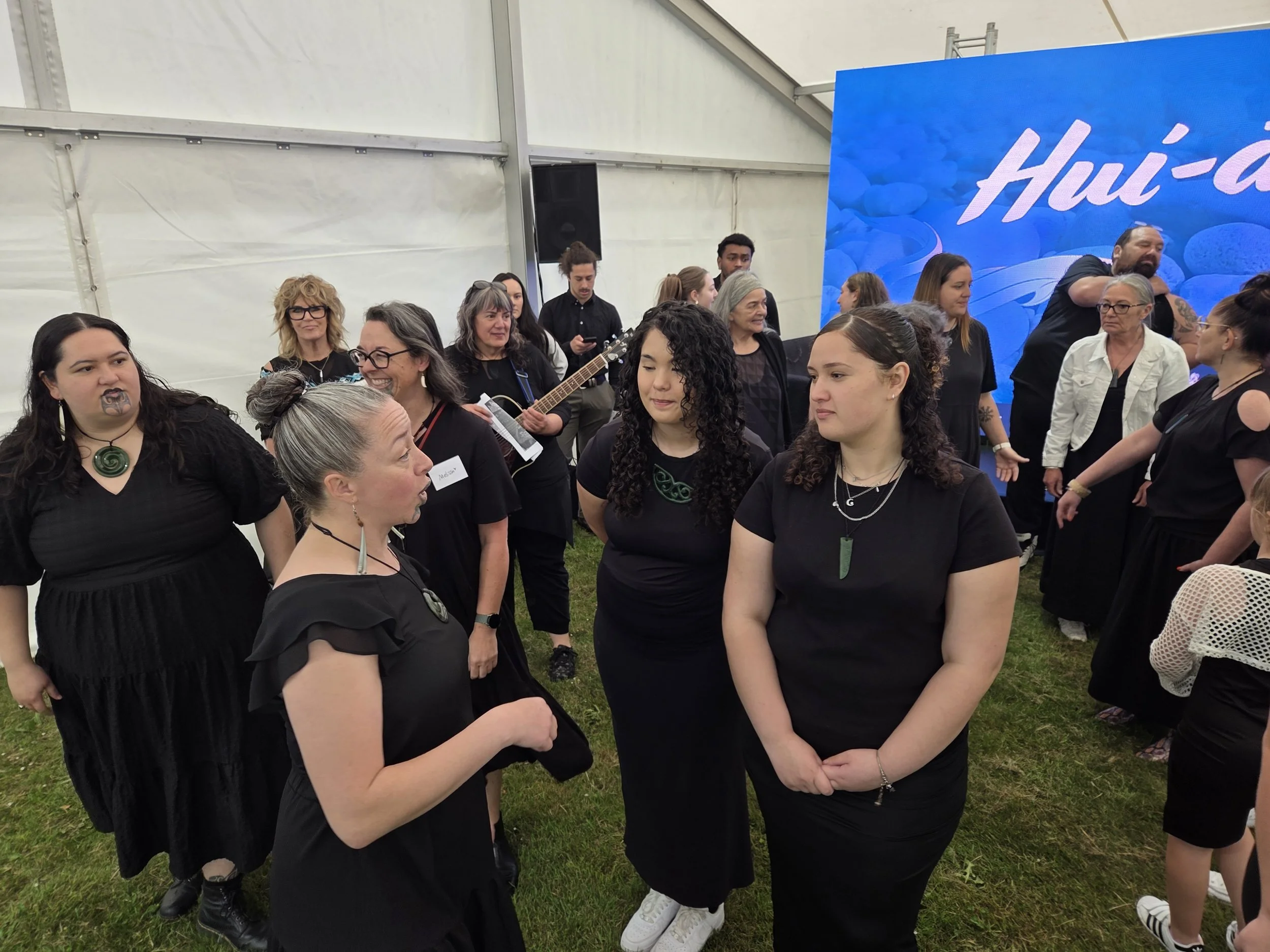 A group of people, mostly women, standing and conversing inside a large tent at an event. Some are holding drinks, and one woman is playing a guitar. In the background, a large screen displays part of a logo or text that reads 'Hui-'. The people appear to be engaged in conversations, wearing dark clothing and accessories, and some are smiling.