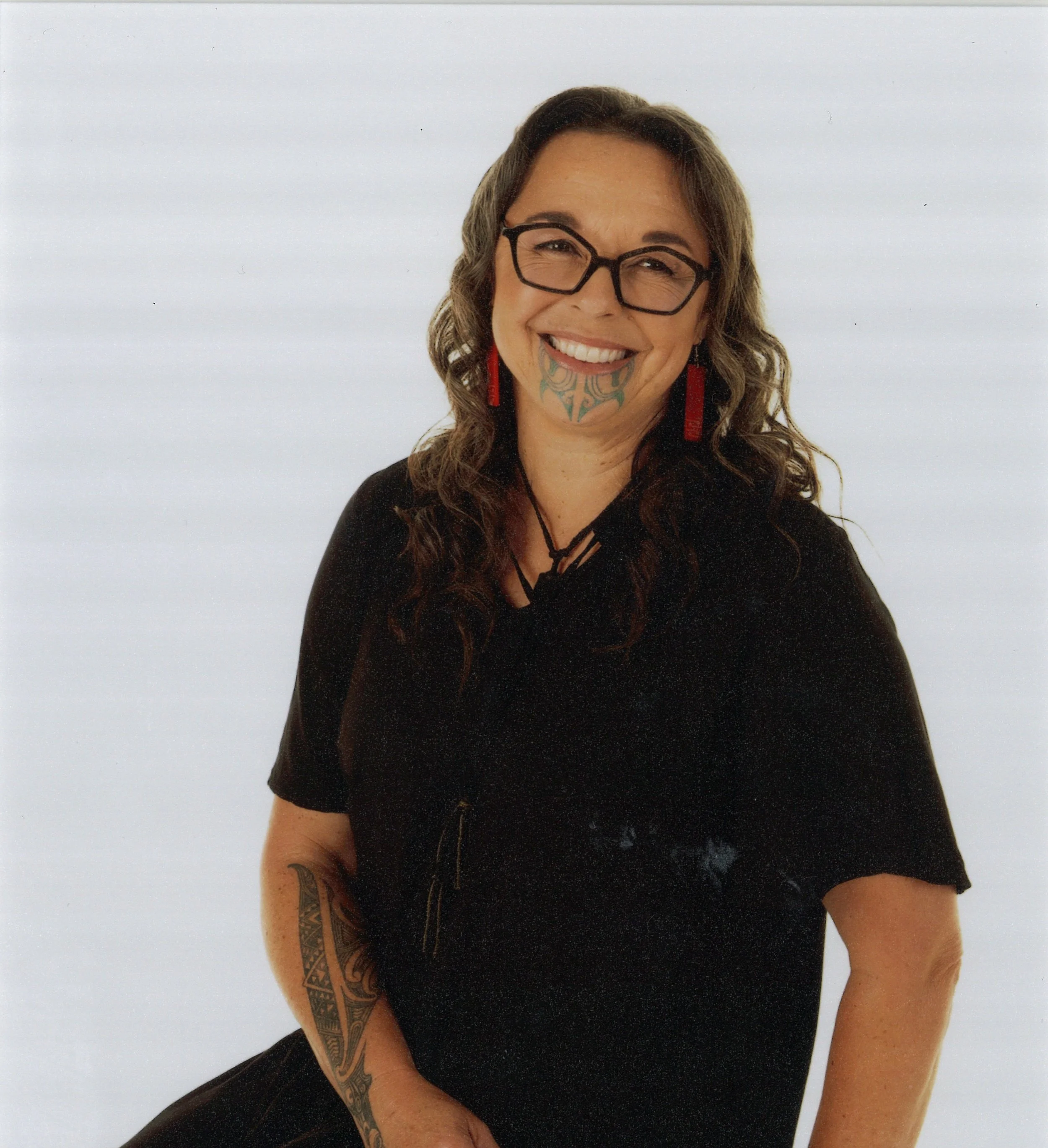 A woman with curly brown hair, wearing glasses and red earrings, smiling, with tattoos on her left arm, wearing a black top, against a plain white background.