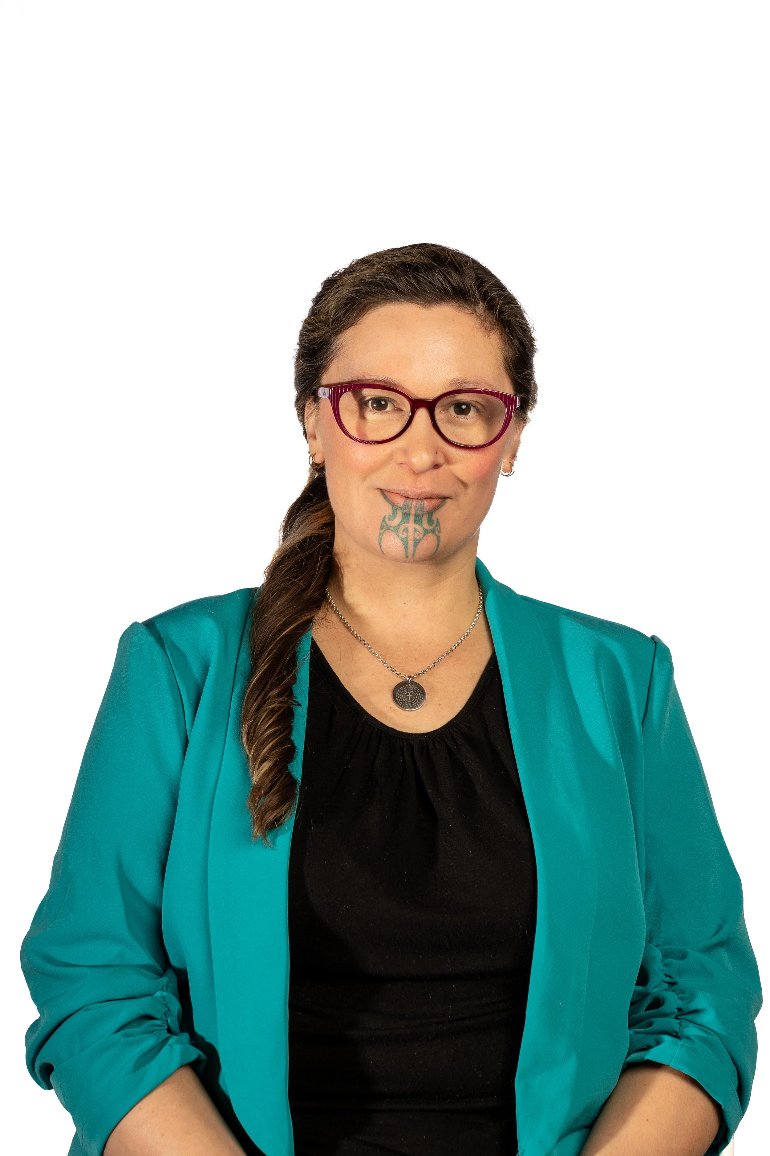 A woman with brown hair in a braid, wearing glasses, a black top, and a teal blazer, with tattoos on her chin and ear, seated against a white background.