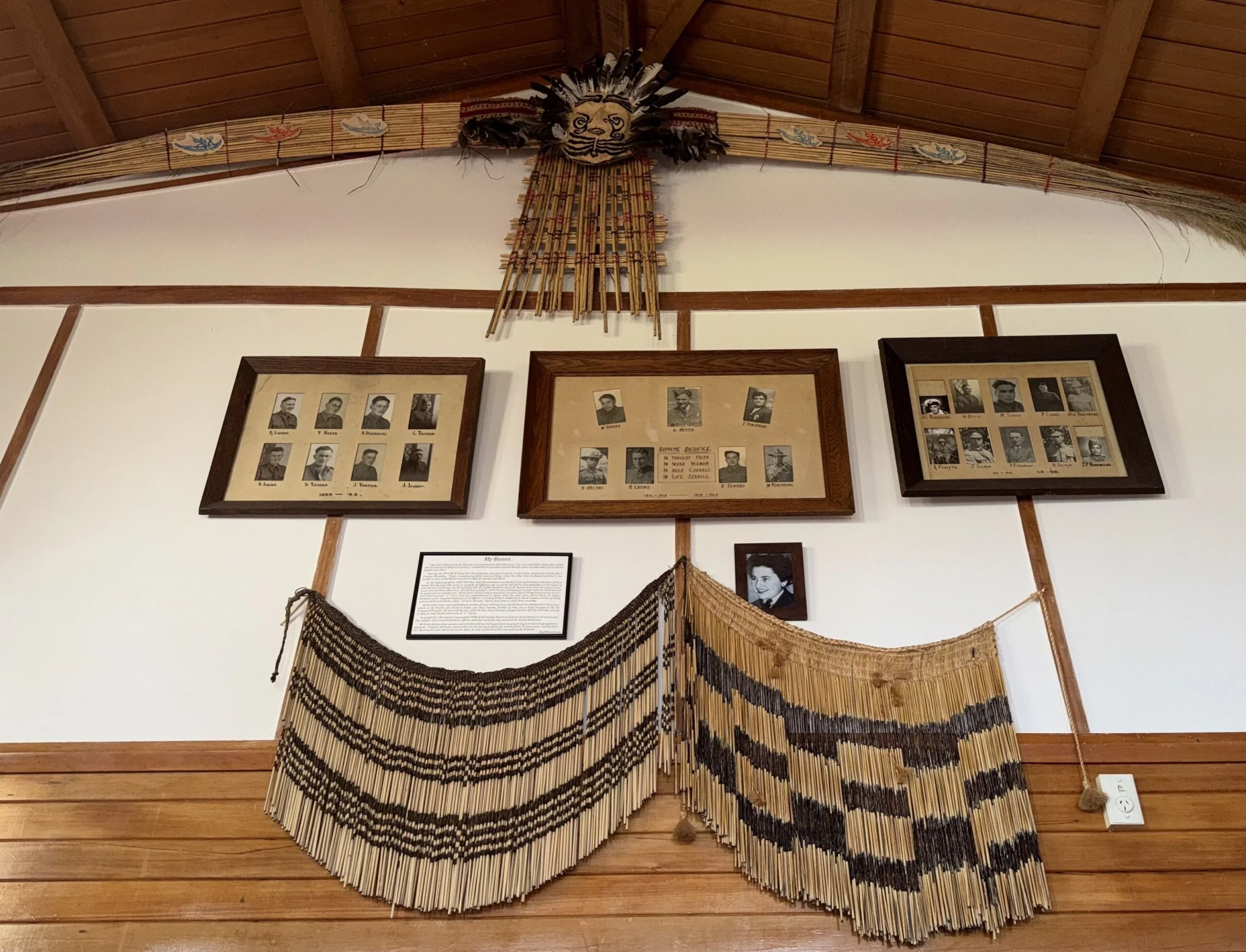 Decorative wall hanging made of beads with a feathered headdress mascot at the top, on a wall with framed black-and-white photographs.