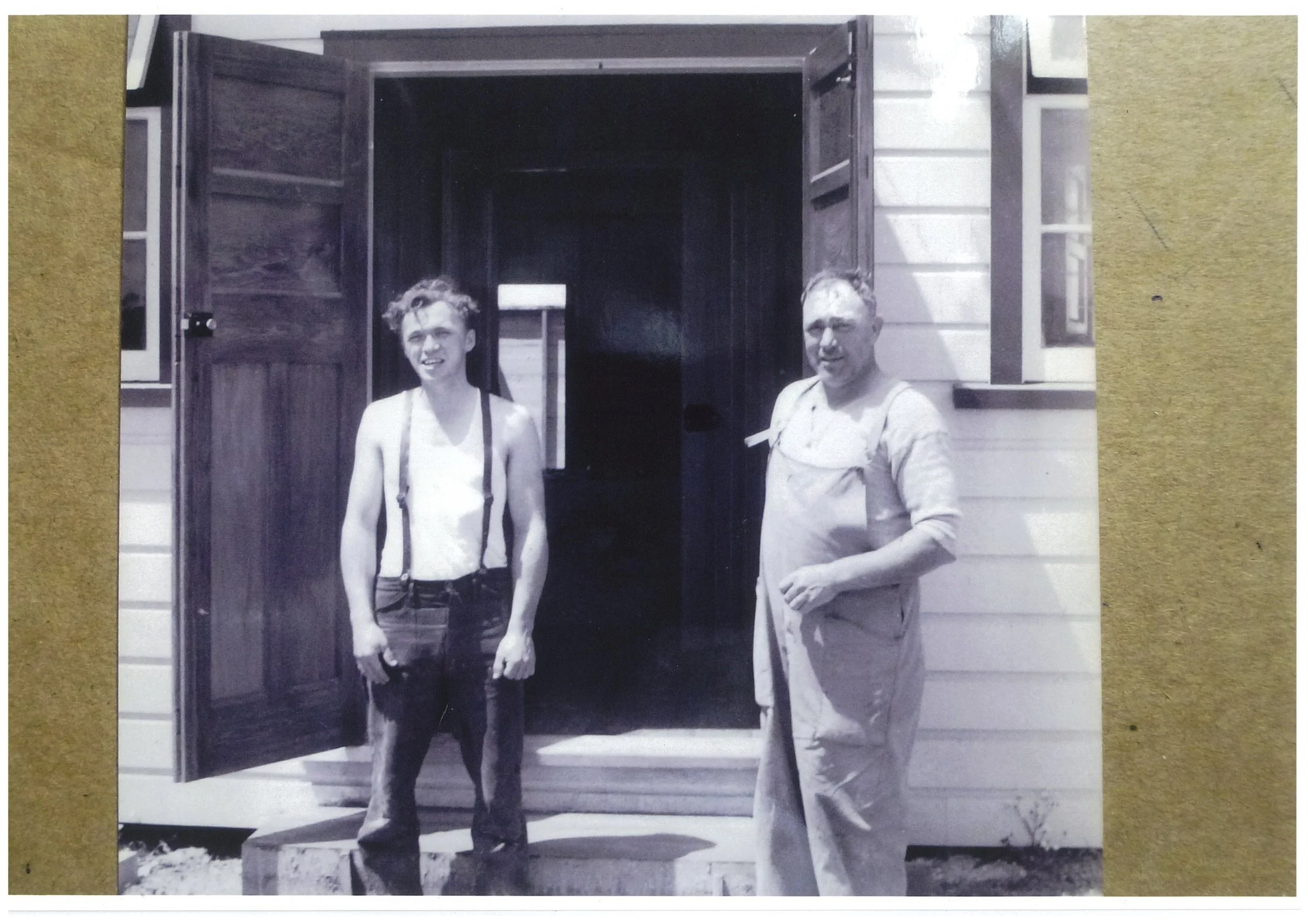 Black-and-white photo of two young men standing in front of a house with open dark wooden door and clapboard siding. One man is shirtless with suspenders, the other is wearing glasses and a light shirt. They are smiling and looking at the camera.