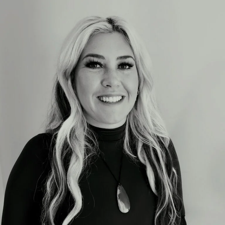 Black and white portrait of a smiling woman with long wavy hair wearing a black top and a pendant necklace.