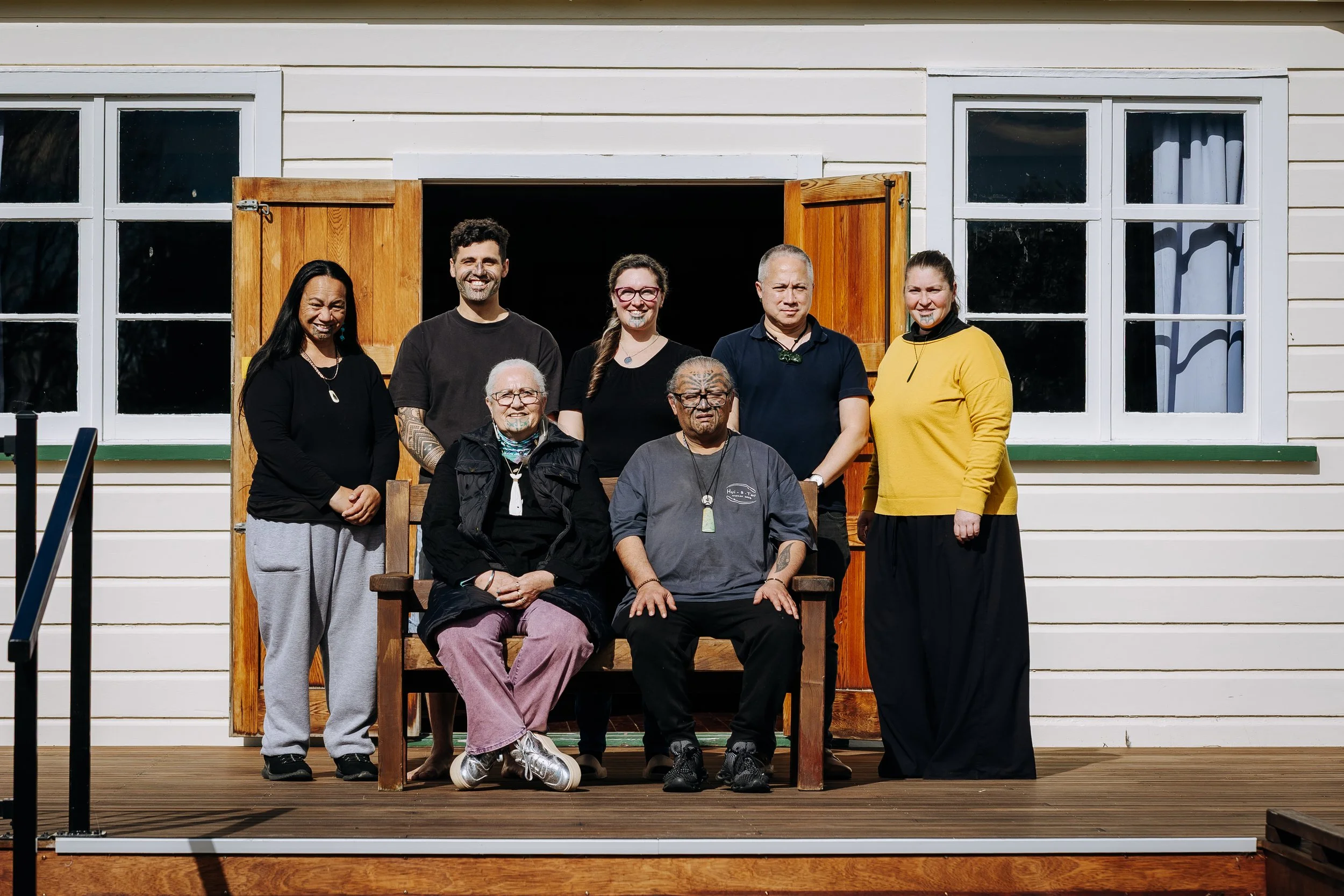 Group of seven people standing and sitting on a wooden porch in front of a white house with two large windows and open wooden door. The group includes diverse individuals with different ages, appearances, and clothing styles, some smiling, others more serious.
